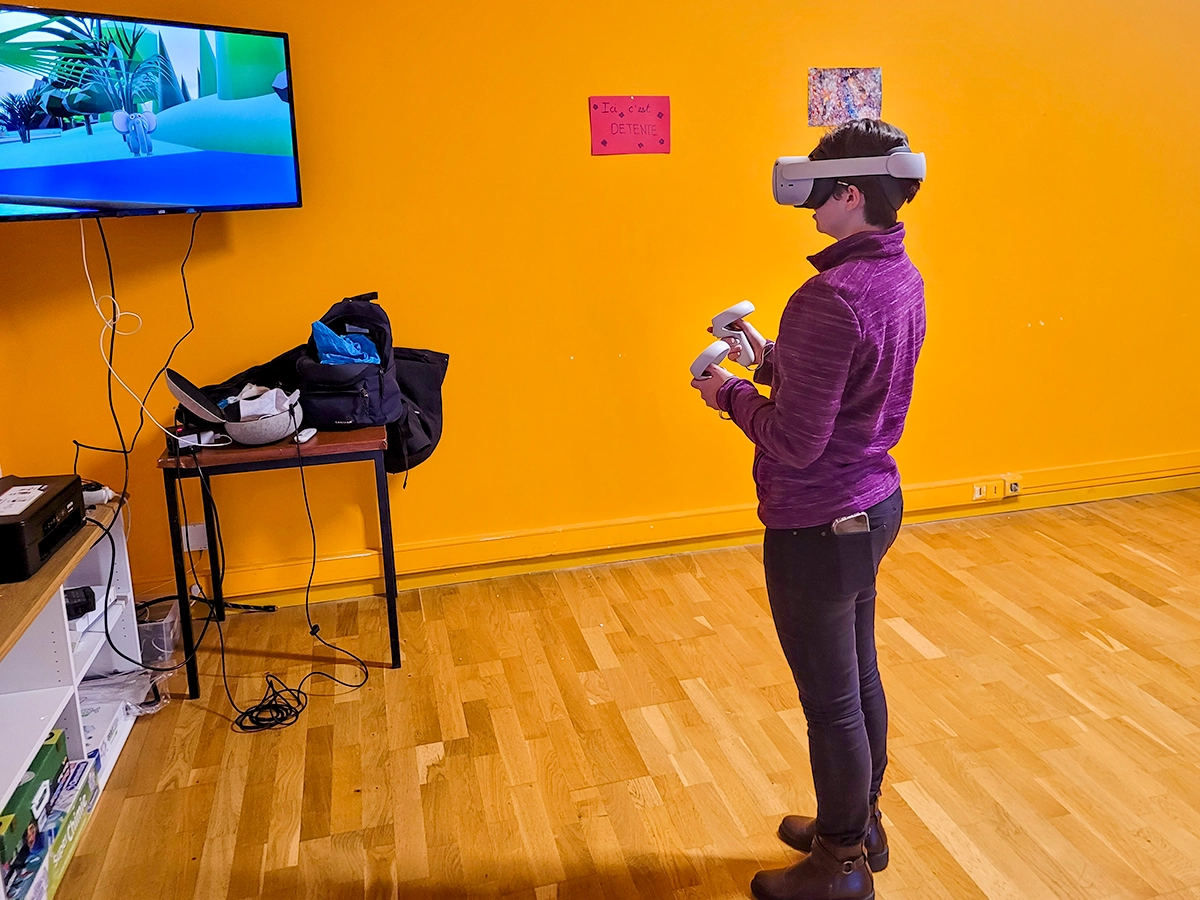 Une personne vue de profil en veste violette utilise un casque de réalité virtuelle et des manettes de contrôle face à un mur orange, un écran diffusant des images de nature virtuelle en arrière-plan.